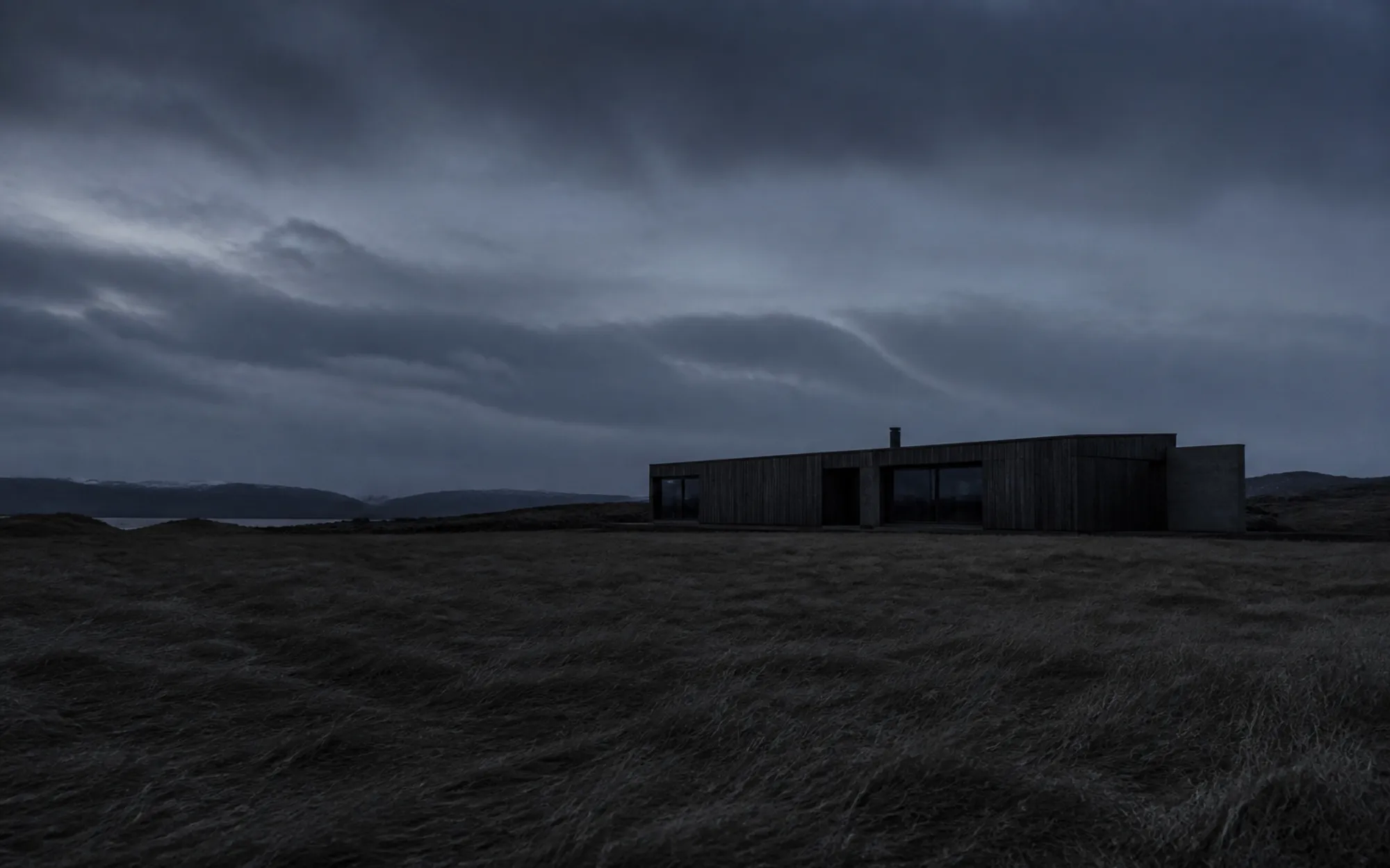 A long low dark-timber building sitting in an open northern landscape at dusk with wind-flattened grass