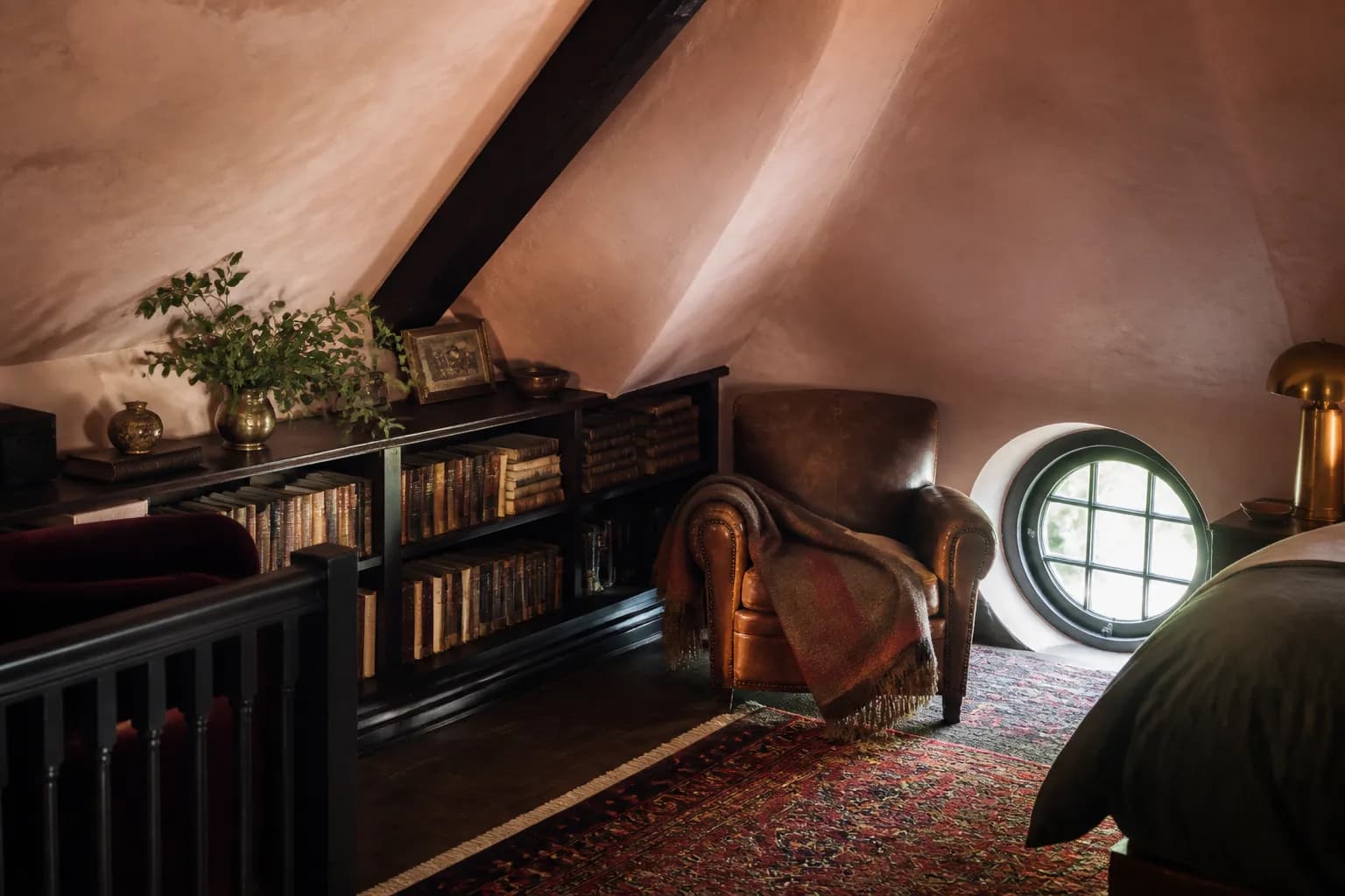 A knee-wall bookcase built into the slope of the roof, a worn leather armchair beside it and a small round window above.