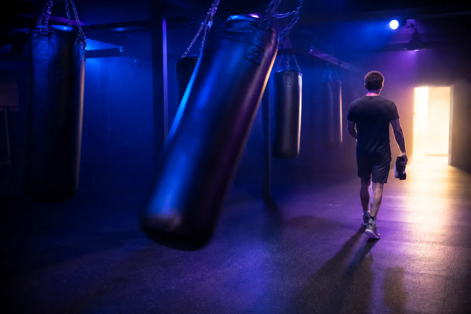 A single heavy bag still swinging in an emptying gym as one person walks toward the lit exit