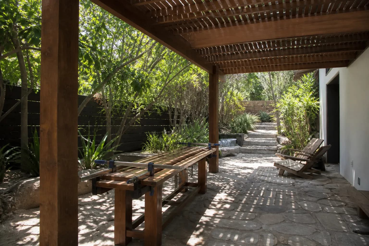 A shaded patio under a simple wood pergola, a level and clamps still lying on the bench below.