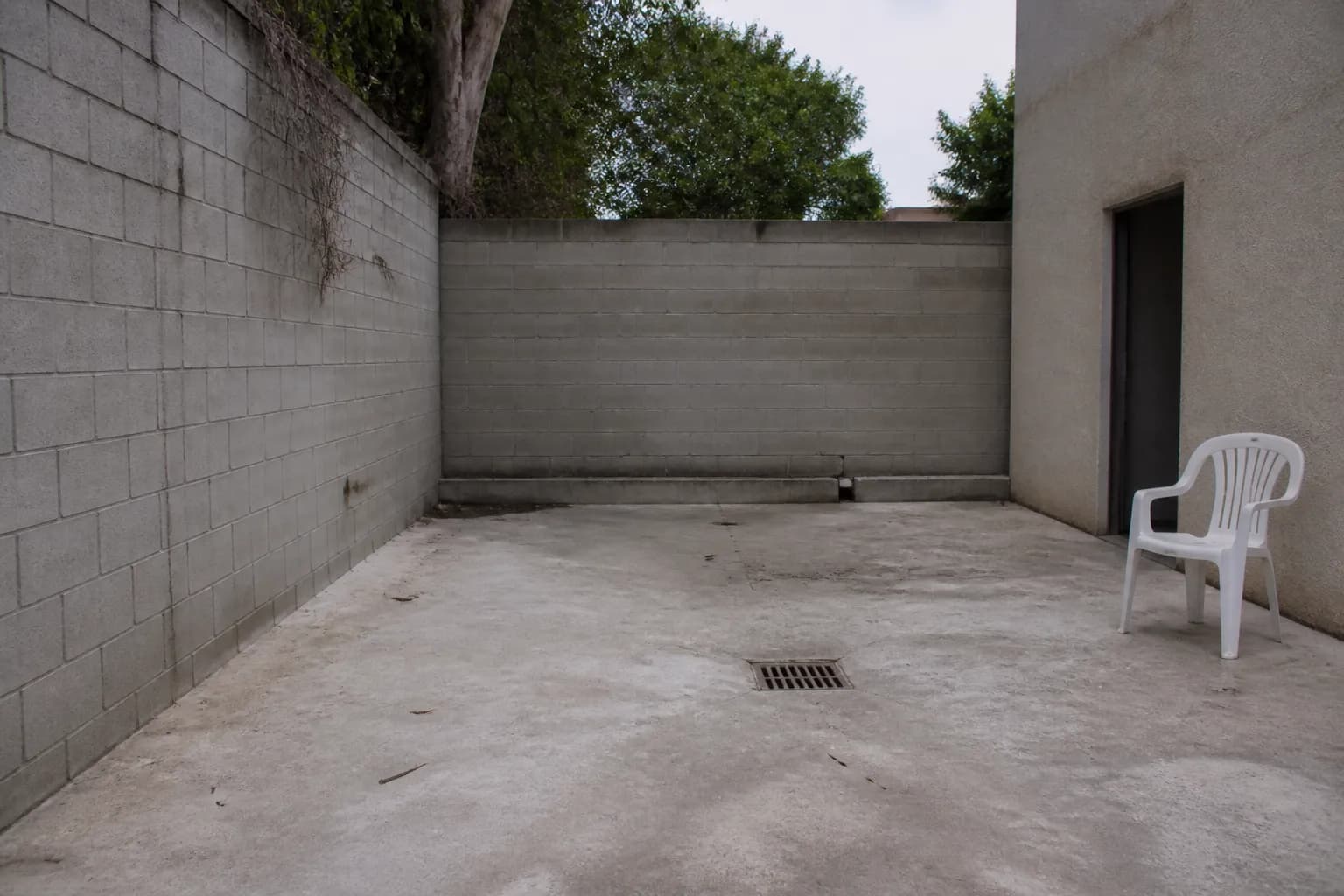 A plain concrete courtyard with bare walls, a floor drain and a single plastic chair in flat light.