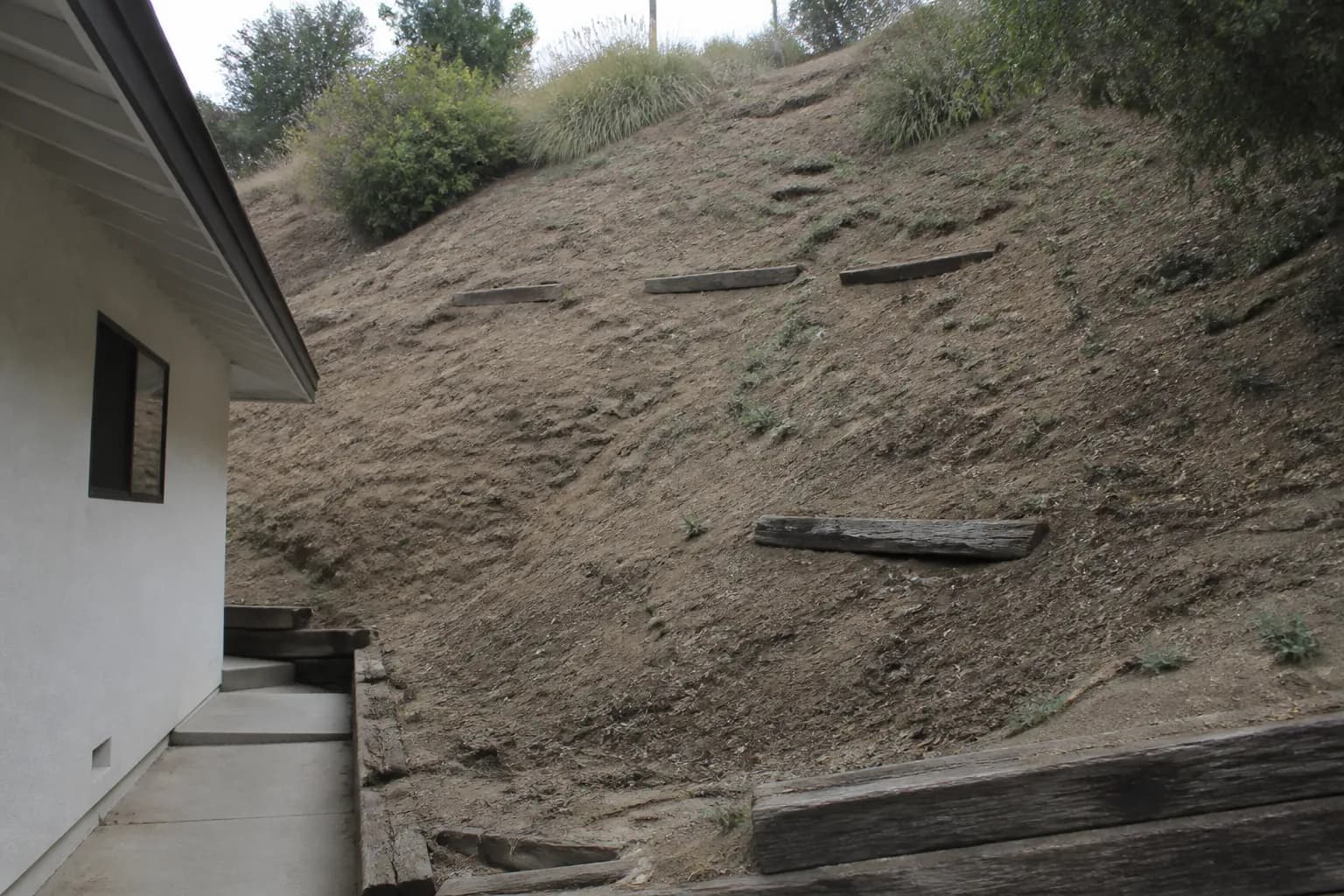 A steep bare slope behind a house with exposed dry soil, a few weeds and failing railroad ties.