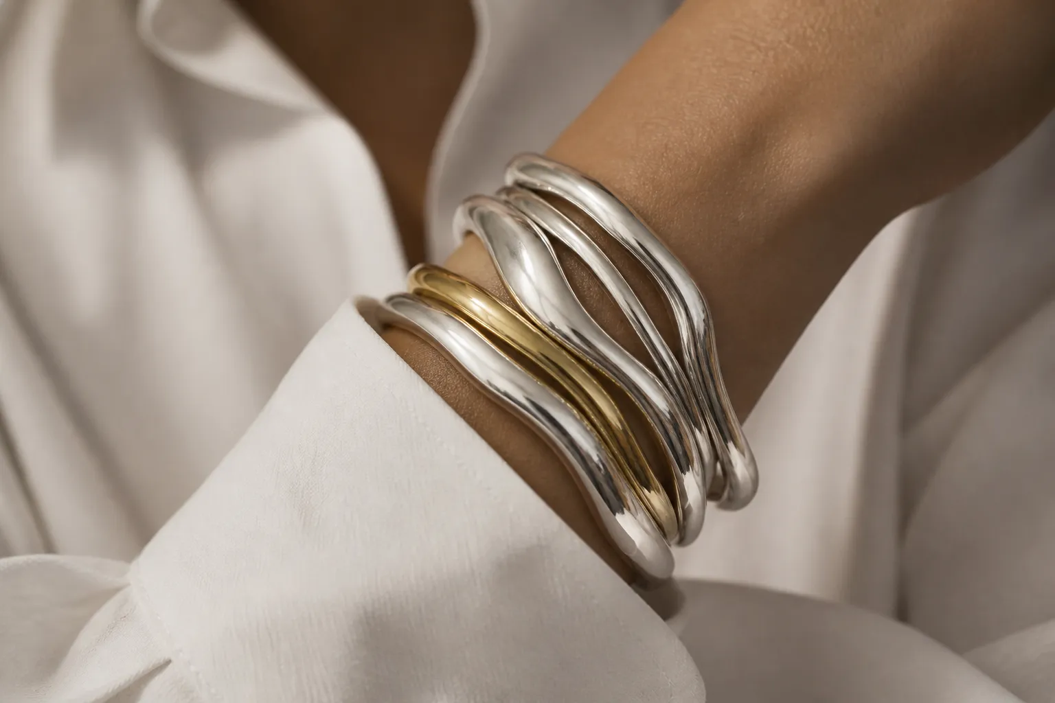 A wrist crossed over a white cotton shirt cuff wearing a stack of sculptural silver bangles with one gold bangle among them.