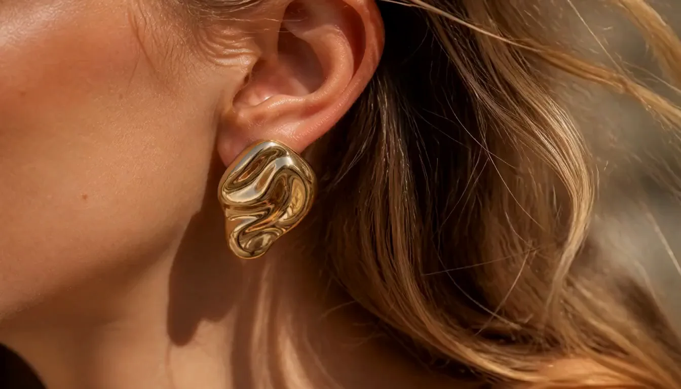 Macro photograph of an ear and jaw line wearing a thick fluid gold sculptural earring, with loose dark blonde hair catching warm daylight.