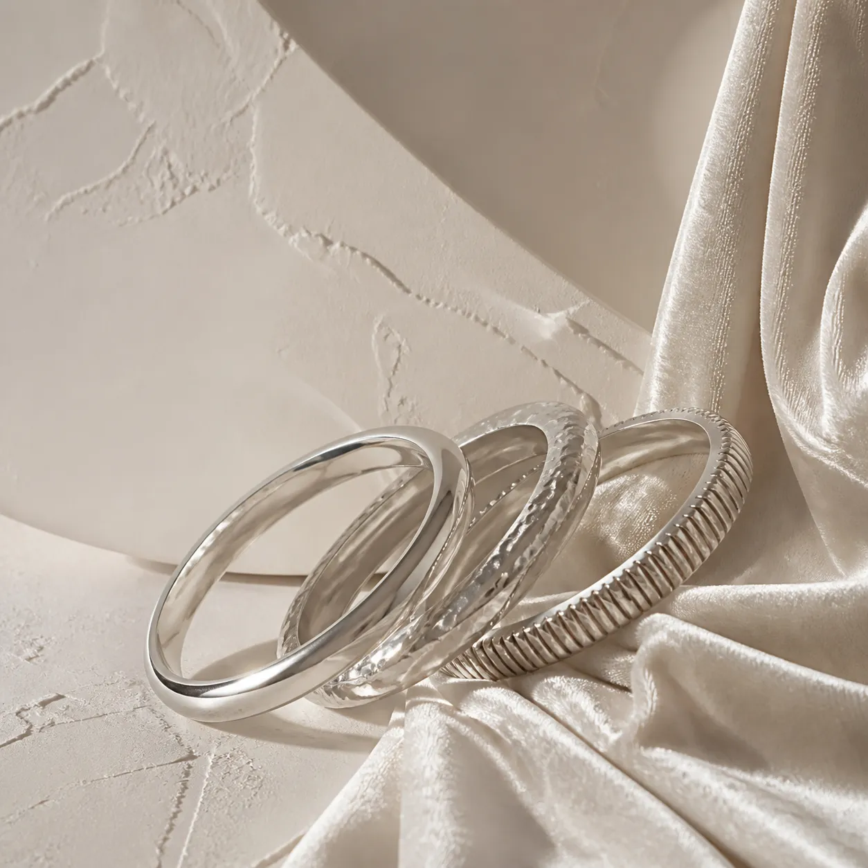 Three sculptural silver bangles leaning against each other on ivory velvet.