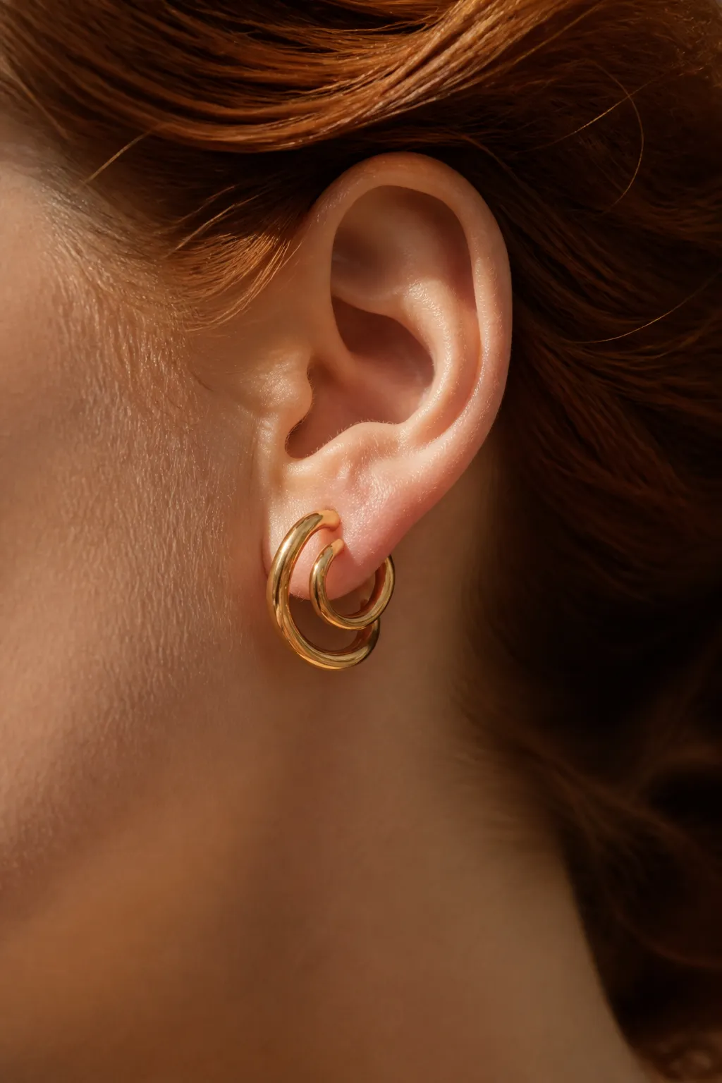 An ear with swept-back auburn hair wearing a fluid gold hoop that wraps the lobe twice.