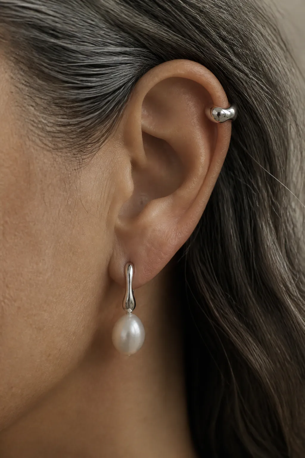 An ear with grey-streaked hair tucked behind it, wearing a small silver cuff high on the helix and a drop below.