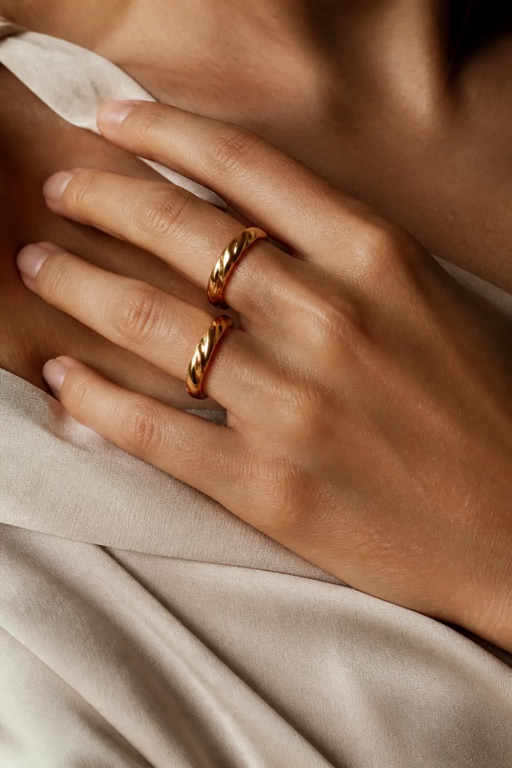 A hand resting on a collarbone wearing two twisted gold rings, soft fabric texture below.