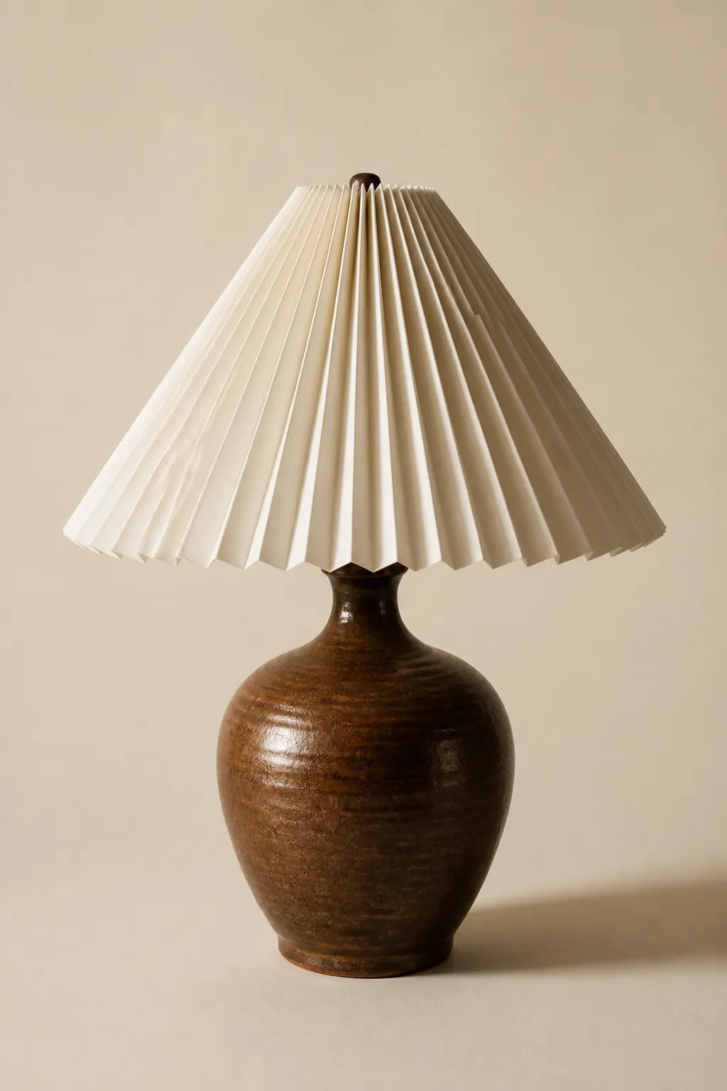 A sculptural table lamp with a hand-thrown ceramic base in umber glaze and a pleated paper shade, photographed on a warm cream background.
