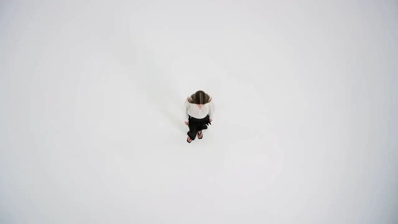 Seen from directly above, a model in a white ruffle blouse and black flared trousers stands on a bright studio floor